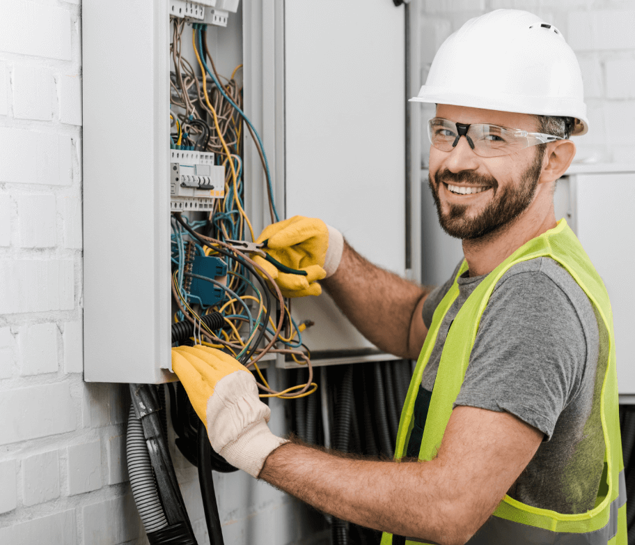 A male electrician smiling at the camera, whilst at work wearing a hard hat and high vis jacket